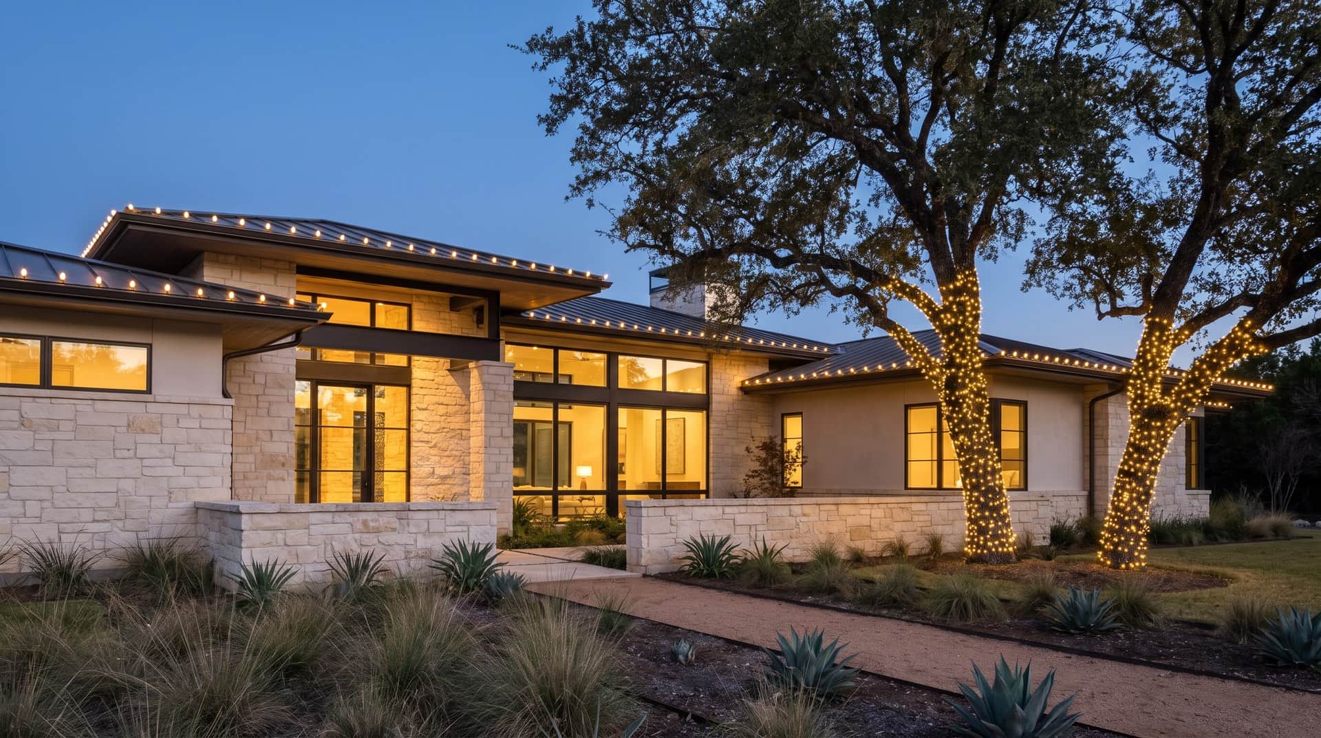Design mockup: a Hill Country contemporary home of limestone, stucco and steel with a low-slope metal roof under large live oaks in West Austin at dusk, with warm white lines of light on the roof edges and wrapped oak trunks