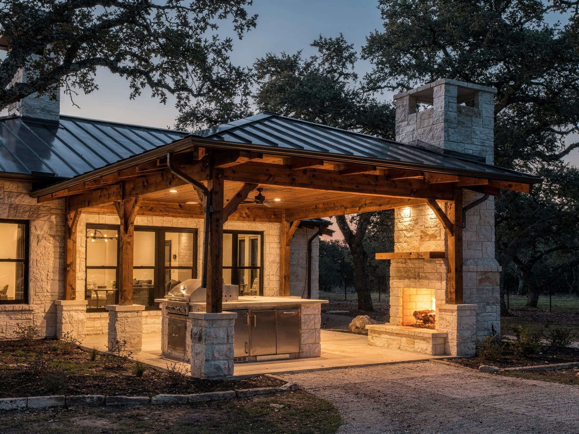 Metal-roof patio cover over an outdoor kitchen and stone fireplace at dusk