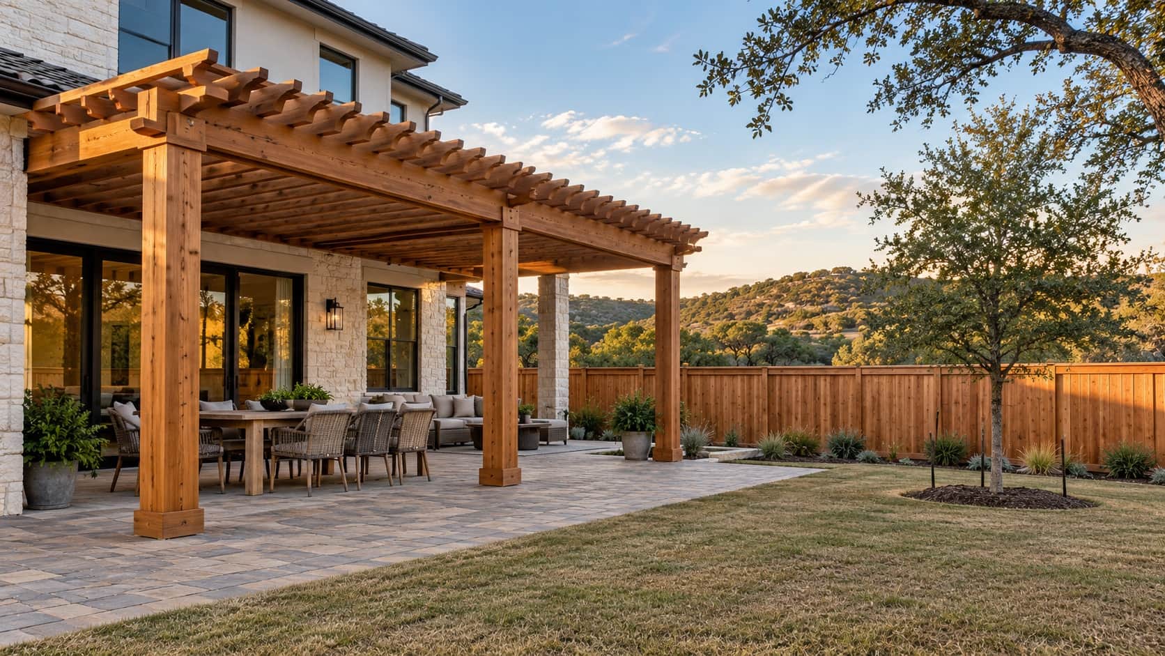 Stained cedar pergola over a paver patio behind a newer stone-and-stucco home with a live oak and Hill Country sky, the kind Klema builds in Esperanza