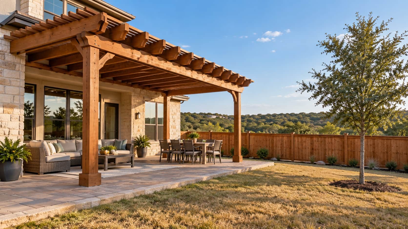 Stained cedar pergola over a paver patio behind a newer stone-and-stucco home with a cedar privacy fence and a young live oak, the kind Klema builds in Kinder Ranch