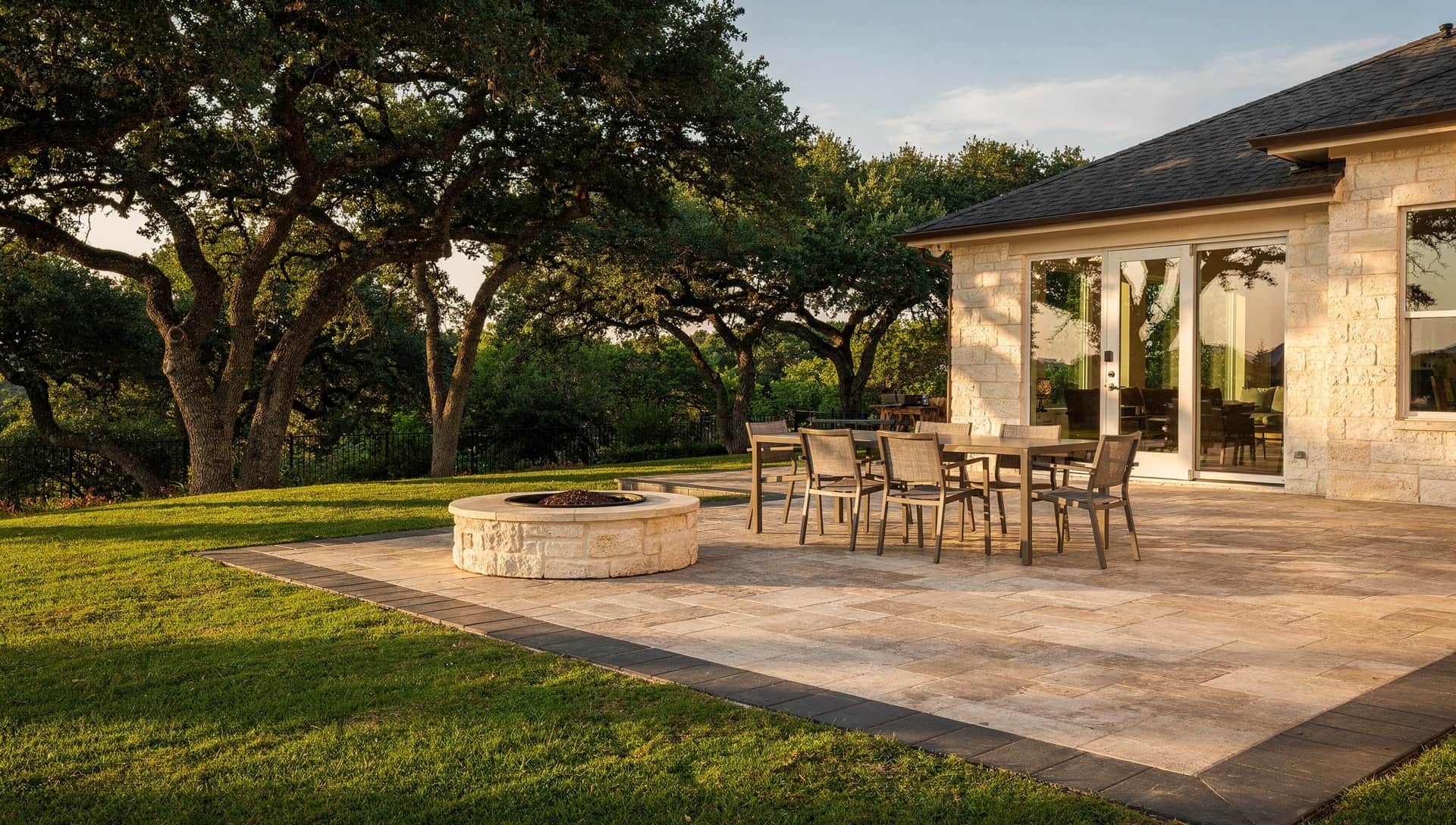 Tan paver patio with a charcoal border, a dining table and a limestone fire pit behind a Hill Country home