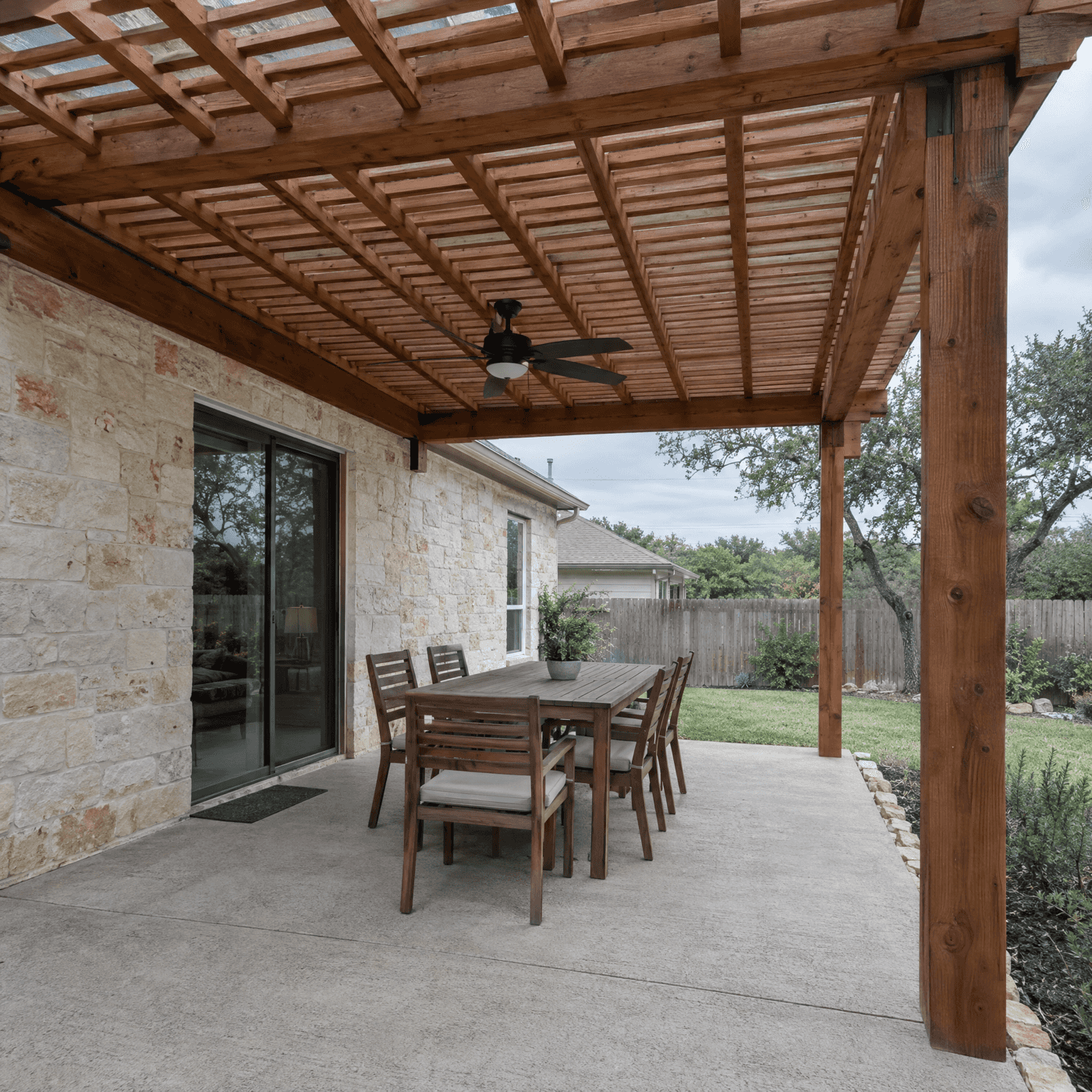 Cedar pergola attached to a limestone home with a ceiling fan over a concrete patio